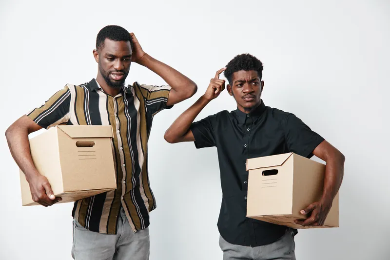 Two young black men with dark skin holding cardboard boxes and scratching heads in confusion or doubt on white background. Casual clothing and uncertain expressions.