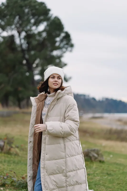 Outdoor woman wearing a long beige puffer coat in a chilly landscape with trees and distant shoreline, showcasing casual winter fashion and warm, comfortable style.