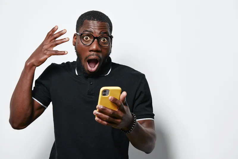Black man with dark skin and glasses holding yellow smartphone with shocked expression and raised hand on white background. Casual clothing and surprised reaction to phone content.