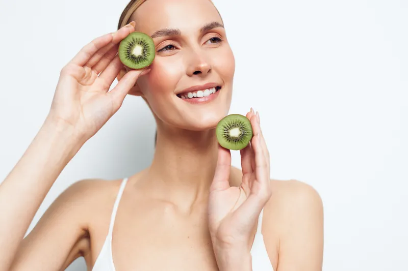 A smiling young woman in a white top holds two halves of fresh kiwi fruit near her face against a white background. Concept for healthy lifestyle, natural beauty, and wellness.