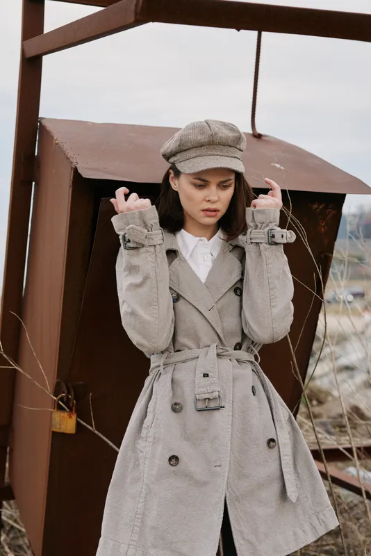 Young woman in tweed coat and matching cap stands outdoors near rusty metal structure, looking down with serious expression. Autumn fashion concept with vintage style and textured fabric details.