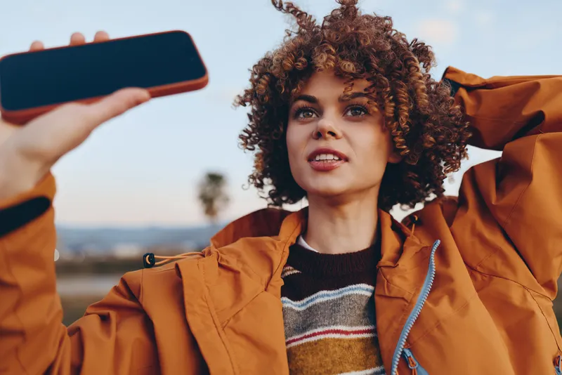 Woman lifestyle selfie outdoors portrait, smartphone in hand, curly hair woman in orange jacket, bright sky and distant hills behind, casual travel mood, content creation concept today