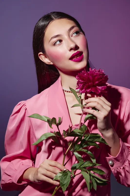 Young woman with glossy brown hair wearing pink blouse and vibrant makeup holding a red flower. Studio shot on purple background. Beauty, fashion, creative concept.