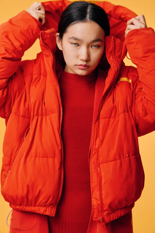 Young woman wearing bright orange puffer jacket and matching sweater standing against yellow background, adjusting hood with focused expression in studio portrait fashion photography.