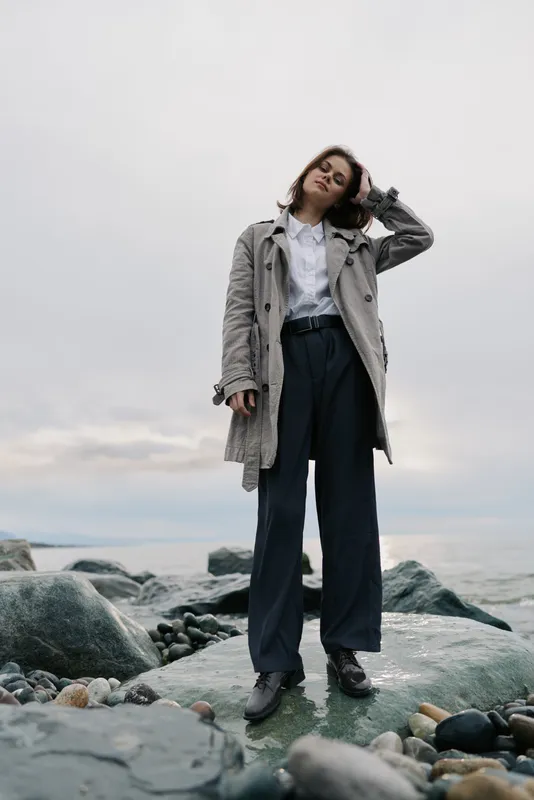 Young woman in stylish coat and wide trousers stands on rocky shore by the sea under cloudy sky. Fashion concept with casual outfit and natural outdoor scenery for autumn or spring.