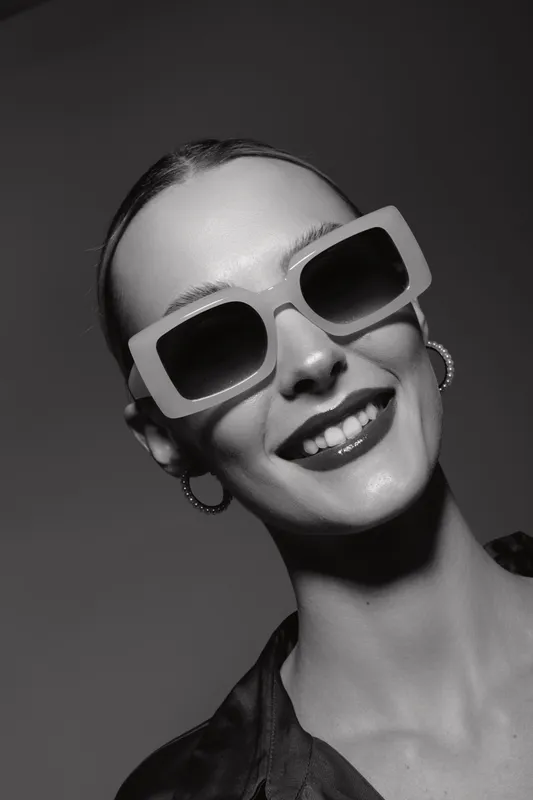 Young white woman with fair skin smiles wearing large square sunglasses and hoop earrings in black and white portrait on grey background.
