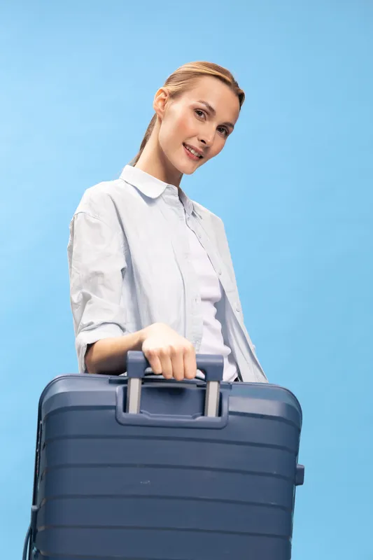 Smiling woman holding blue suitcase with handle, dressed in casual white shirt, standing against blue background. Concept of travel, vacation, and trip preparation.
