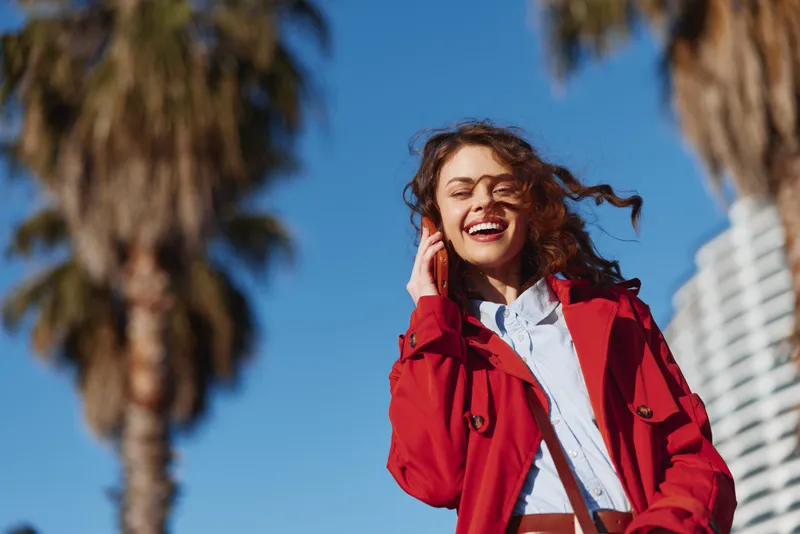 Smiling woman in a red coat poses outdoors on a sunny day, blue sky and palm trees behind. Energetic, casual portrait expressing confidence and fashion.