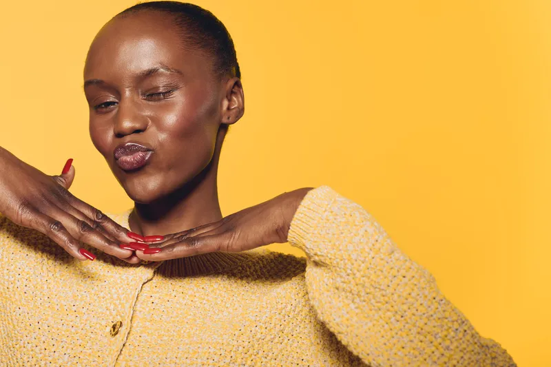 Young African woman with dark skin poses with hands under chin and puckered lips, wearing yellow sweater against yellow background. Concept of beauty, confidence, and self-expression.