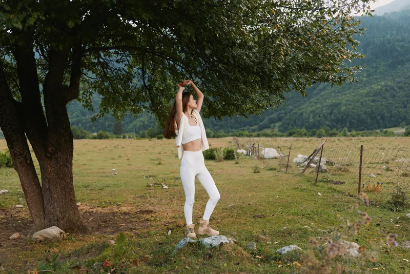 Woman in white dances under a tree in a sunlit meadow, enjoying carefree lifestyle moments in a rural landscape, outdoor happiness