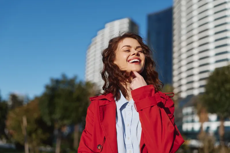 Smiling woman outdoors in a red jacket, urban cityscape behind, bright sunlight enhances a casual portrait with confident modern fashion vibe and energetic vibe