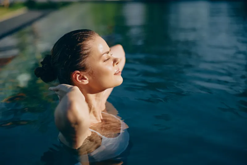 Young woman with brunette hair relaxes in an outdoor swimming pool with her eyes closed and hands behind her head enjoying a peaceful moment on a sunny day.