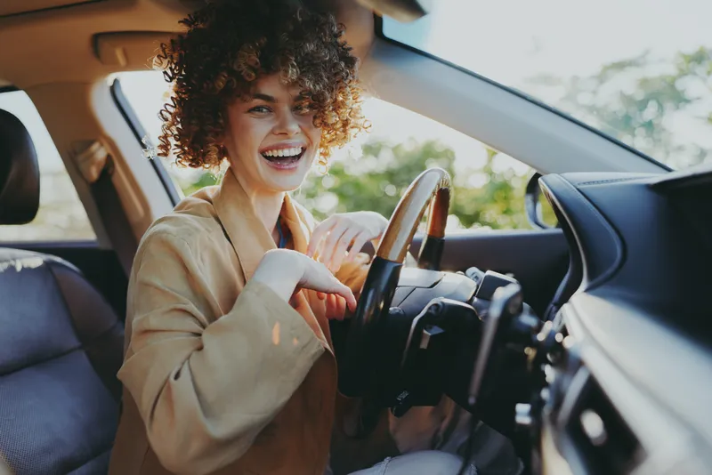 Smiling woman with curly hair driving a car, casual jacket, daylight streaming through the window. Clear lifestyle moment focusing on travel, independence, and everyday transportation.