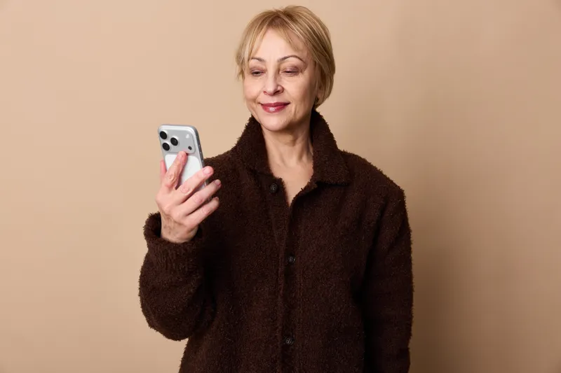 Smiling woman holding smartphone in portrait, wearing winter coat, using mobile communication technology, standing against beige studio background, copy space