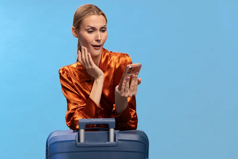 Curious woman in orange satin blouse looks surprised at smartphone screen while leaning on blue suitcase. Concept of travel, mobile communication, and unexpected news on blue background.