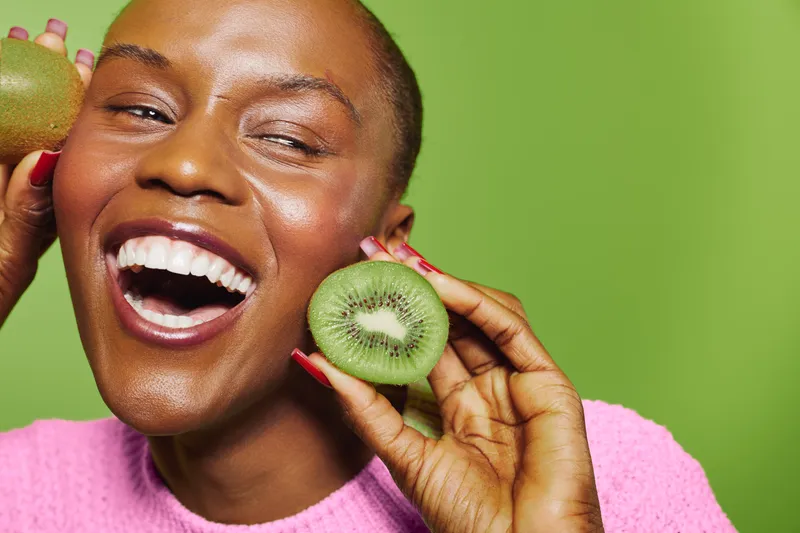 Happy African woman with dark skin holds fresh kiwi fruit near face, smiling broadly in front of green background. Concept of healthy lifestyle, fruit, wellness, and natural beauty.