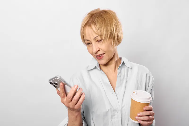 Elderly woman using smartphone indoors, holding a paper coffee cup, gentle smile, focused gaze at screen, relaxed posture against plain white background, casual day