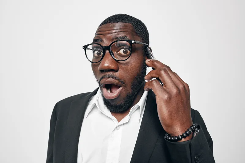 African American man with dark skin and glasses showing surprised expression while talking on smartphone in formal suit against white background.