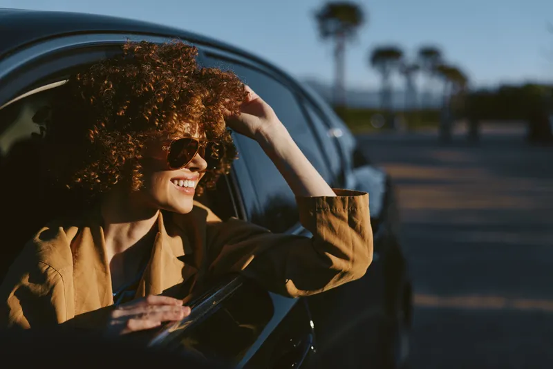 A woman with curly hair in a tan jacket smiles while leaning out of a car window, sunlit street scene with retro film color and a carefree lifestyle vibe.