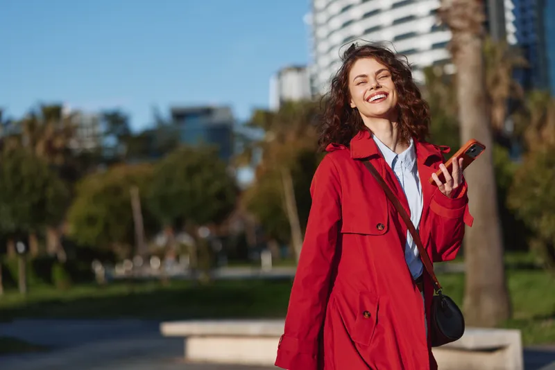 Smiling woman with curly hair wearing red coat outdoors holding smartphone in sunny urban park. Concept of happiness, technology, and lifestyle in casual fashion.
