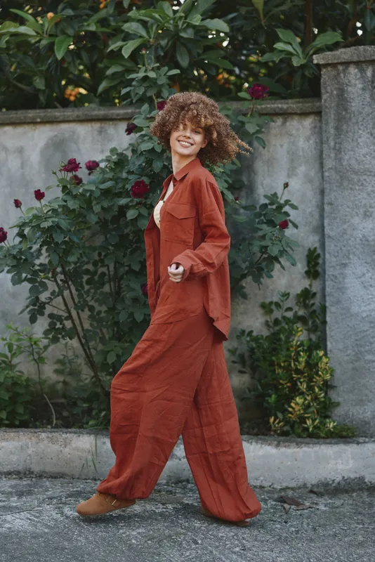 Smiling woman with curly hair walking outdoors in a rust jumpsuit, garden roses in the background, sunny street mood, casual fashion, confident lifestyle