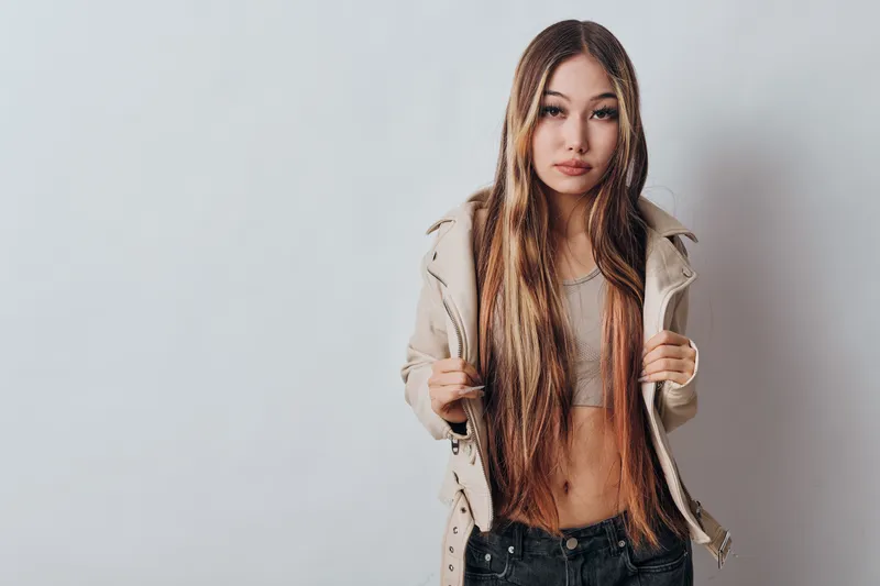 Young woman with long straight hair wearing a beige jacket and crop top posing against a plain grey background with confident expression and casual style.