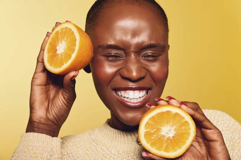 Happy African woman with dark skin holding two orange halves near her face smiles with eyes closed on yellow background. Concept of healthy lifestyle, vitamin, and natural beauty.