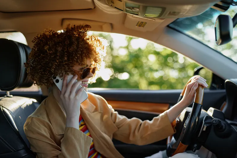 A stylish woman driver with curly hair and sunglasses sits behind the wheel in a sunlit car interior, showcasing retro fashion and everyday travel mindset.