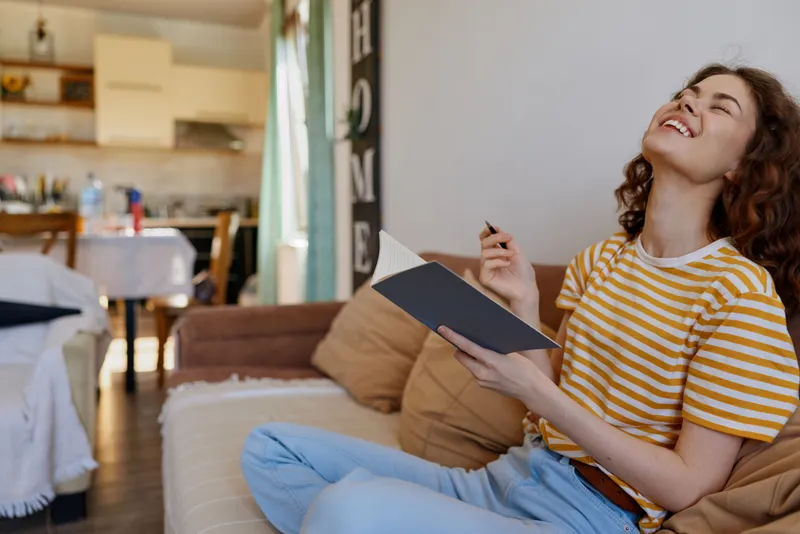 A joyful woman sits on a sofa at home, reading a notebook and smiling, with bright natural light filling the room. A cozy lifestyle moment centered on comfort.