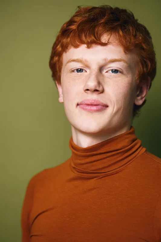 Portrait of young man with red hair and fair skin wearing burnt orange turtleneck looking confident with slight smile and neutral green background in studio lighting.