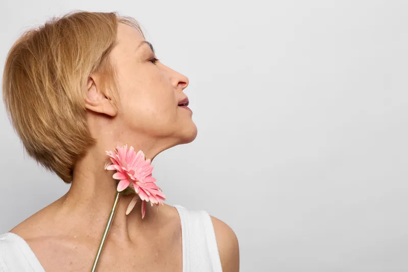 Woman flower closeup profile studio portrait spring pink gerbera blow, adult woman in white top smelling petals, soft studio light, isolated neutral background, copy space
