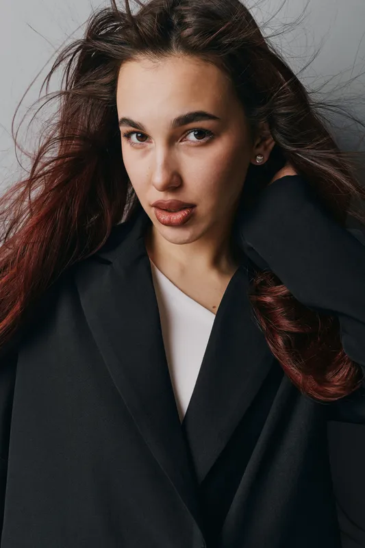 Woman with long brown hair wearing black coat and white top posing with hand in hair against dark background. Portrait of confident female with natural makeup and earrings.