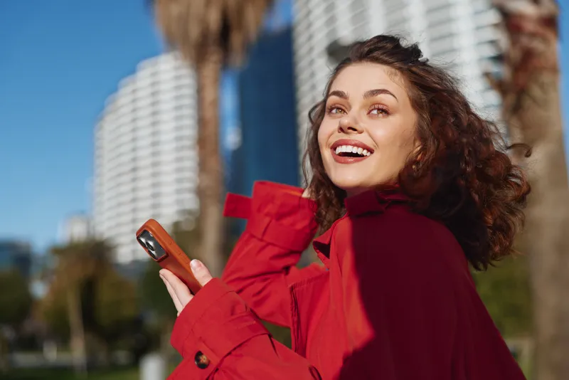 Young woman with curly hair smiles joyfully while holding a smartphone outdoors in a red jacket on a sunny day with urban buildings in the background.