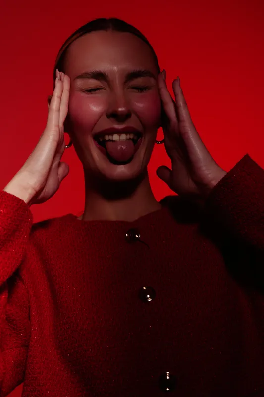 Young woman in red sweater poses with eyes closed and tongue out, hands on head. Studio portrait with red background and playful, expressive mood.