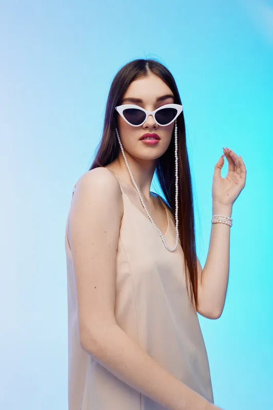 Young woman with long hair wearing white sunglasses and a beige dress poses against a blue gradient background. Concept of beauty, fashion, and modern style.