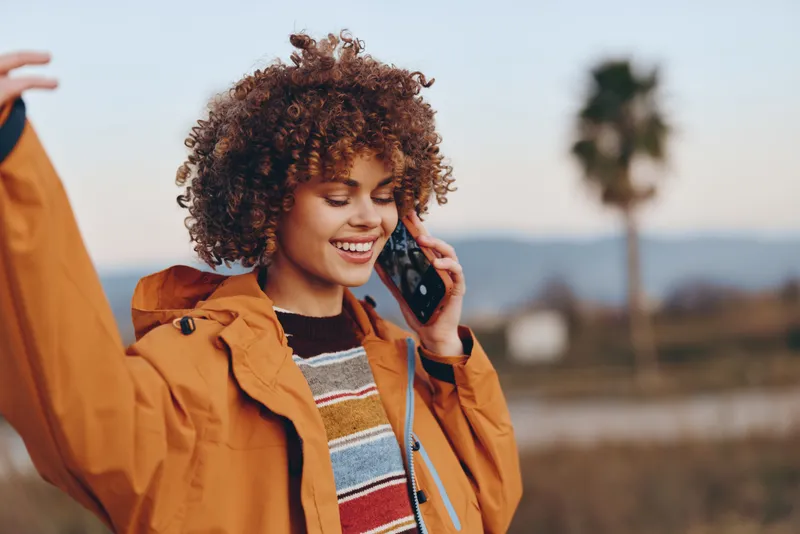 Smiling woman with curly hair in a brown jacket, talking on phone outdoors. Soft golden light and distant landscape create casual lifestyle mood, friendly expression, and shallow depth.