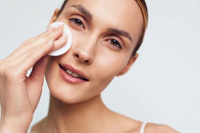 Young woman with clear skin gently removes makeup using a cotton pad on a white background, expressing calmness and self-care in a bright studio setting.