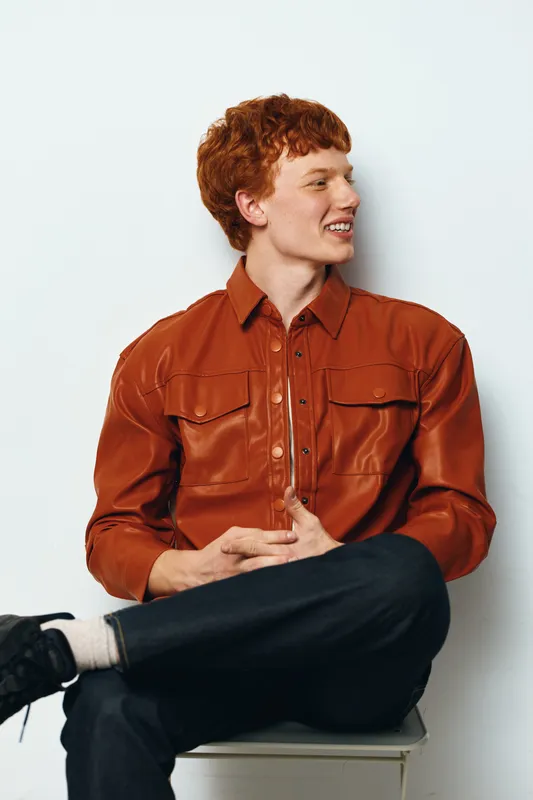 Young man with red hair sitting on chair looking sideways smiling, wearing brown leather shirt and black pants in casual studio portrait on white background.