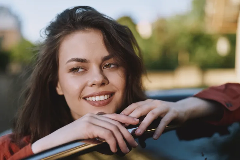 Woman lifestyle portrait smiling in natural light, leaning on a car window, relaxed casual vibe, outdoor urban setting, candid joyful moment