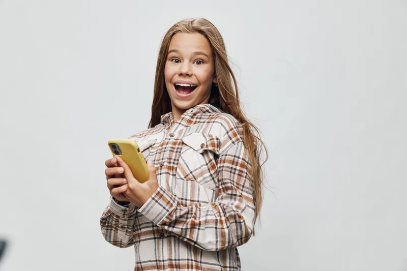 Girl in plaid shirt holding yellow smartphone looking excited and surprised with open mouth on gray background. Teenager and digital device for communication and social media.