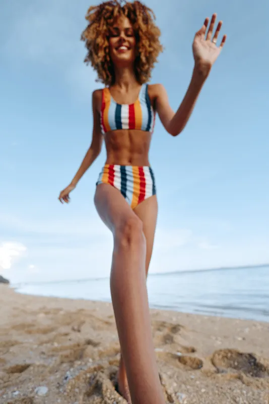 Woman beach ocean bikini summer waving curly hair happiness smiling standing on sandy shoreline near ocean waves under blue sky bright sunlight carefree holiday mood portrait full body