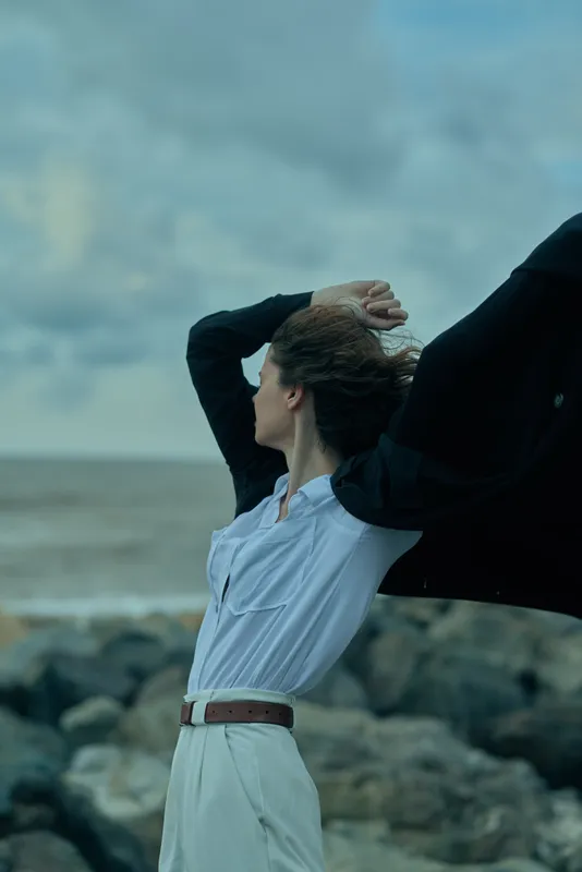 Wind sweeps a flowing cape and white shirt as a figure stands on a rocky coast, conveying freedom, dynamic movement, and bold fashion against a moody sky.