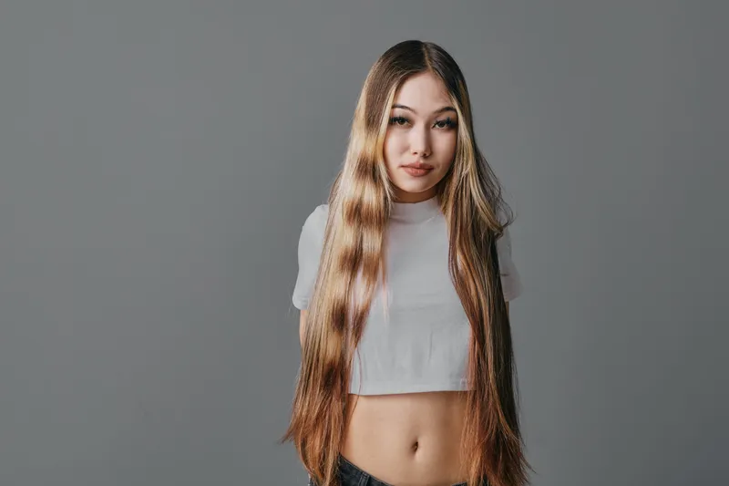 Woman with long hair wearing a white crop top standing against a gray background. Portrait of confident young female with casual style and natural makeup in simple studio setting.