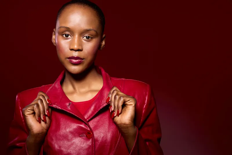 Confident African woman with dark skin wearing a red leather jacket poses against a dark red background. Studio portrait captures strength, fashion, and elegance with vibrant colors.