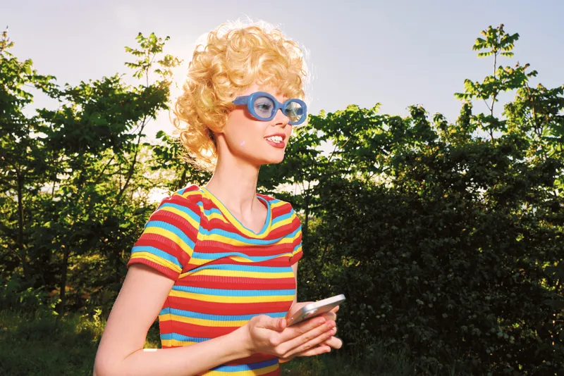 Cheerful woman outdoors, wearing a striped shirt and retro sunglasses, sunlit moment, relaxed posture, soft breeze, natural colors, lush greenery background, with warm glow and mood