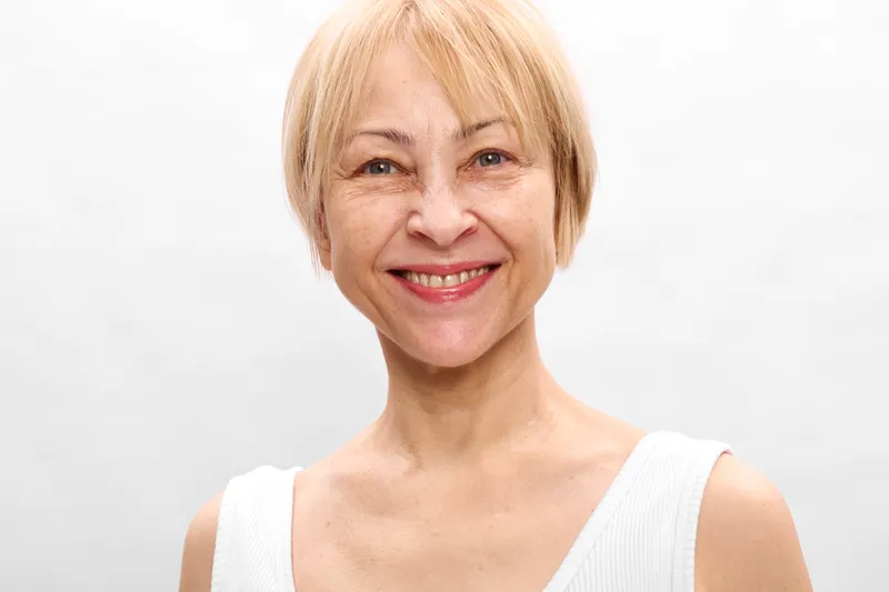 elderly woman smiling portrait of a senior blonde woman with short hair in a white sleeveless top, neutral studio background, bright friendly expression, confidence and wellness, skincare theme