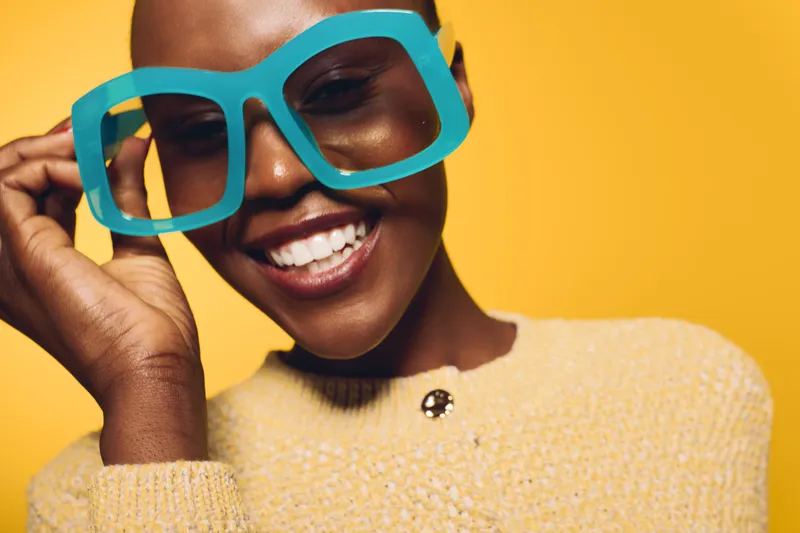 Close-up of a happy black woman with dark skin wearing oversized blue glasses and a yellow sweater. Bright yellow background creates cheerful and playful mood, concept of fashion and joy.