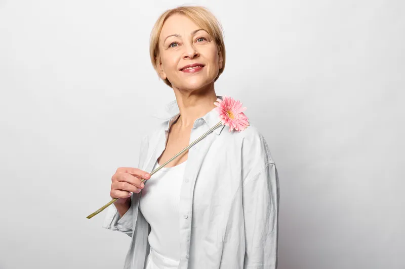 Elderly nurse doctor woman in white coat holds stethoscope and pink carnation flower, smiling in clean studio background, healthcare and medicine concept for gentle care wellbeing