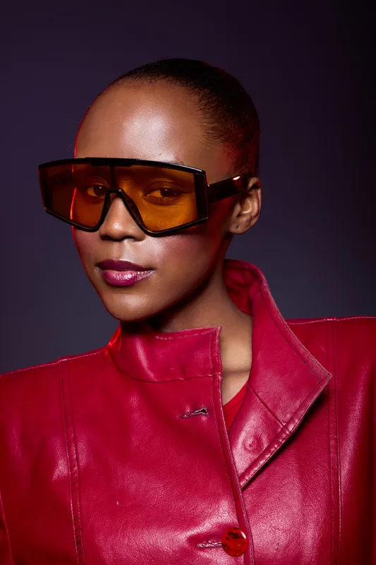 Portrait of a confident black woman with dark skin wearing large orange sunglasses and a red leather jacket. Studio shot with dramatic lighting and stylish fashion concept.