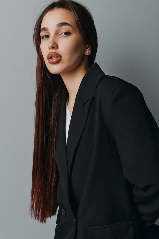 Woman with long straight hair wearing black blazer posing against gray wall in studio. Portrait of confident female model with natural makeup and serious expression looking at camera.